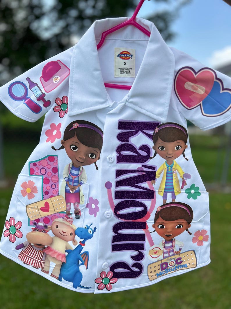 Op de afbeelding: Witte laboratoriumjas voor kinderen met Doc McStuffins-personage. De jas heeft een paarse glitter naam, karakterafbeeldingen en medische illustraties. Een roze hanger ondersteunt de jas.