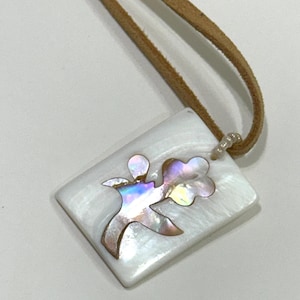 Puede incluir: Un colgante rectangular blanco con un diseño floral de nácar iridiscente. El colgante está unido a un cordón de cuero marrón.