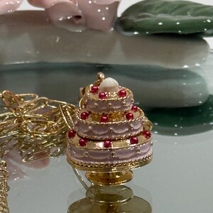 May include: A gold-colored necklace with a three-tiered cake pendant. The pendant is pink with red accents and a white top. The cake sits on a gold stand. The necklace is on a reflective surface.