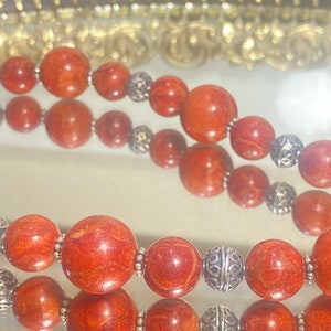 May include: Close-up of two strands of coral bead necklaces. The beads are a vibrant orange-red color, interspersed with ornate silver-colored beads. The necklaces are displayed on a reflective surface, with a blurred decorative background.