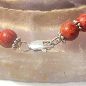 May include: Close-up of a coral bead necklace with a silver clasp. The beads are round and a reddish-orange color. The clasp is a silver lobster claw style. The necklace is on a light-colored surface.