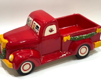 May include: A red ceramic pickup truck with a decorative design. The truck features a Christmas theme with a wreath, snowman, and other festive details. The wheels are black with white and red accents.