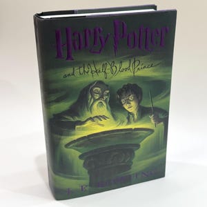 Puede incluir: Un libro de tapa dura verde y negro con el título "Harry Potter y el Príncipe Mestizo" en letras moradas. La portada presenta una ilustración de Harry Potter y Albus Dumbledore de pie sobre un caldero brillante.