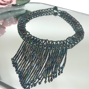 Può includere: Una collana di perline con un design a frange. La collana presenta un colletto di piccole perline scure con una frangia di perline cilindriche più lunghe nei toni del blu, verde e bronzo. La collana è esposta su una superficie riflettente.