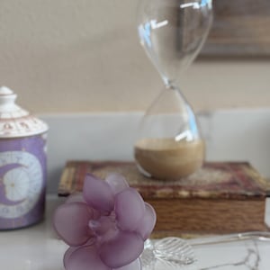 Puede incluir: Un reloj de arena de cristal transparente con arena marrón en su interior se encuentra sobre un libro antiguo. Una flor morada esmerilada y una hoja plateada están en primer plano. Un tarro decorativo morado con tapa blanca está a la izquierda.