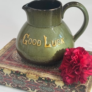 Könnte beinhalten: Eine grüne Keramikkrug mit schwarzem Innenraum und gebogenem Griff. Die Worte "Good Luck" sind in goldfarbenen Buchstaben auf der Vorderseite geprägt. Eine rote Blume liegt auf einem dekorativen Buch.