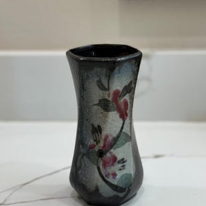 Pode incluir: Um pequeno vaso hexagonal de cerâmica preta com um design floral. O vaso tem um fundo branco com flores e folhas rosa e verdes.