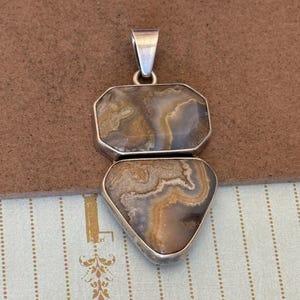 Puede incluir: Un colgante de plata con dos piedras superpuestas de forma irregular con patrones de remolinos en tonos marrón, bronceado, gris y blanco. La piedra superior es rectangular con esquinas redondeadas y la inferior es triangular.