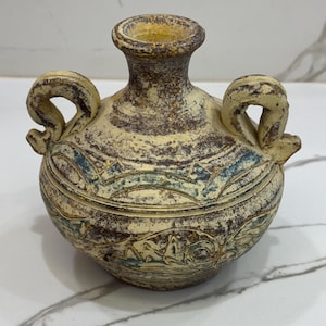 Puede incluir: Un pequeño jarrón de cerámica de color crema con dos asas. El jarrón tiene un diseño marrón y azul con un patrón geométrico.