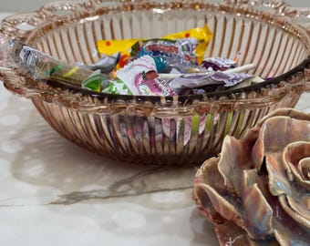 Pode incluir: Uma tigela de vidro rosa com borda ondulada cheia de doces embalados. A tigela tem ranhuras verticais e fica ao lado de uma escultura de rosa de cerâmica com tons rosa, azul e castanho.
