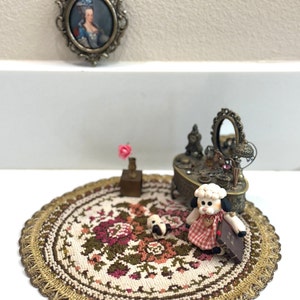 Può includere: Una scena di casa delle bambole in miniatura con una bambola di pecora bianca che indossa un vestito rosso e bianco seduta su un tappeto rotondo floreale. Una piccola statuetta di cane è accanto alla pecora. Un piccolo comodino con uno specchio e un piccolo ritratto incorniciato sono sullo sfondo.