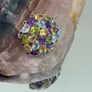 Puede incluir: Un anillo de plata con un conjunto de piedras preciosas ovaladas en varios colores, incluyendo morado, amarillo, verde y azul. El anillo está colocado sobre una superficie texturizada y clara, creando un contraste llamativo. Las piedras preciosas están dispuestas juntas, formando un diseño vibrante y llamativo.