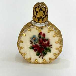 Puede incluir: Un frasco de perfume vintage con un diseño de filigrana dorada. El frasco presenta un diseño floral bordado en punto de cruz en rojo, azul y verde sobre un fondo crema. El frasco es redondo con una tapa dorada decorativa.