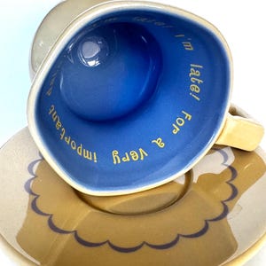 Puede incluir: Un juego de taza y plato de cerámica con exterior color canela e interior azul vibrante. El interior de la taza presenta el texto amarillo "I'm late! I'm late! For a very important date!". El plato tiene un patrón festoneado marrón.