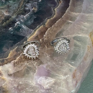 May include: A pair of silver heart-shaped earrings with clear cubic zirconia stones. The earrings are set against a textured, light brown and green background.