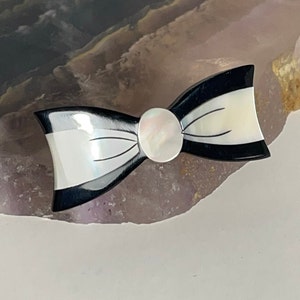 May include: A black and white bow tie brooch with a mother-of-pearl center. The brooch is made of shell and has a polished finish.