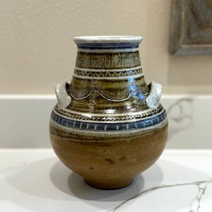 Pode incluir: Um vaso de cerâmica marrom com detalhes em branco e azul. O vaso tem um corpo arredondado e duas pequenas alças nas laterais. Tem um design decorativo de linhas e espirais.