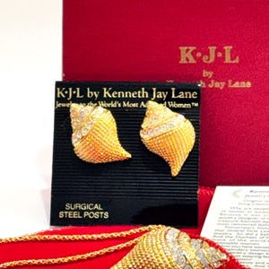 May include: Gold-toned jewelry set by Kenneth Jay Lane, featuring shell-shaped earrings and a pendant necklace. The earrings and pendant are embellished with clear crystals. The set is displayed on a black card with "SURGICAL STEEL POSTS" text and a red velvet pouch.