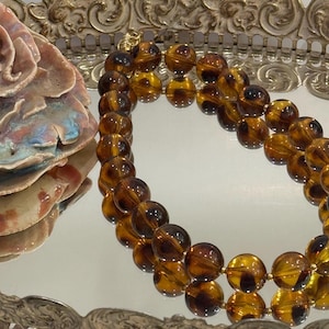 May include: A vintage necklace featuring large, round amber-colored beads. The beads are strung together with gold-toned metal accents. The necklace is displayed on a mirrored surface, reflecting the jewelry and a decorative ceramic piece.