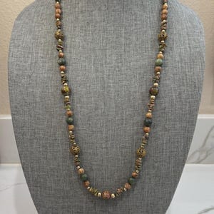 Puede incluir: Un collar largo de cuentas expuesto sobre un maniquí gris. El collar presenta una variedad de cuentas en tonos tierra, incluyendo verde, marrón y dorado, intercaladas con detalles metálicos decorativos. Las cuentas varían en tamaño y forma, creando un aspecto texturizado.