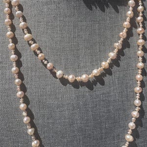 Puede incluir: Un collar largo de múltiples hilos compuesto por numerosas perlas de agua dulce pequeñas, de color rosa pálido y forma irregular. Las perlas varían ligeramente en tamaño y textura, creando un hilo continuo y delicado que cae en bucles.