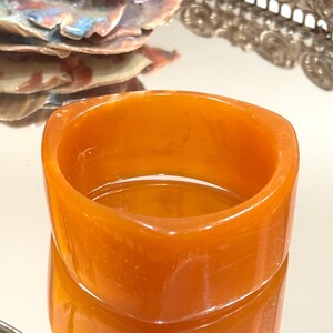 Può includere: Un bracciale a cerchio quadrato e spesso, di colore ambra aranciato. Il bracciale è realizzato in un materiale traslucido che riflette la luce. Il design è semplice e moderno, con una finitura liscia e lucida. Un pezzo audace.