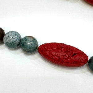 Puede incluir: Collar con cuentas redondas pulidas en tonos verdes, marrones y verde azulado moteado. Incluye una cuenta roja ovalada tallada con intrincados diseños florales, creando una joya única.
