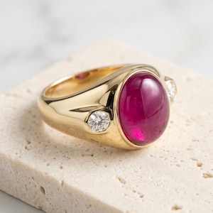 Puede incluir: Un anillo de oro con una gran piedra preciosa cabujón de color rosa intenso y forma ovalada en el centro. La banda es de oro pulido e incluye dos pequeñas piedras redondas y transparentes talladas a cada lado de la piedra central.