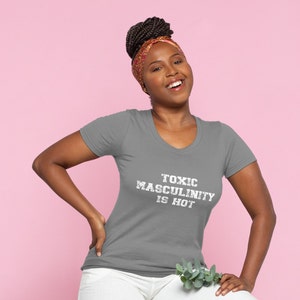 Puede incluir: Una camiseta gris de cuello en V con el texto "Toxic Masculinity is Hot" impreso en blanco en la parte delantera. La camiseta la lleva puesta una persona con el pelo oscuro y una diadema rosa.