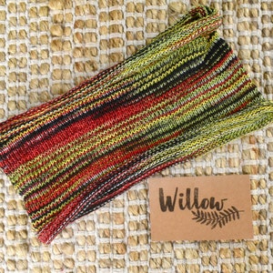 May include: A multi-colored striped knitted headband in shades of red, green, black, and white. The headband is on a woven straw background with a brown tag that says "Willow" with a stylized fern leaf.