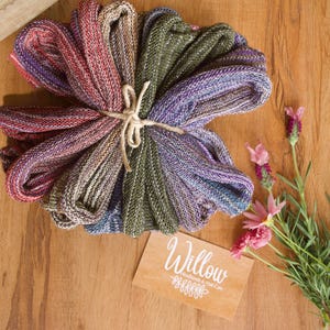 Op de afbeelding: Een bundel kleurrijke gebreide haarbanden, vastgebonden met touw, met tinten paars, groen en rood. Een houten kaart met het woord "Willow" en een bloemmotief ligt naast de haarbanden, met roze bloemen op een houten oppervlak.