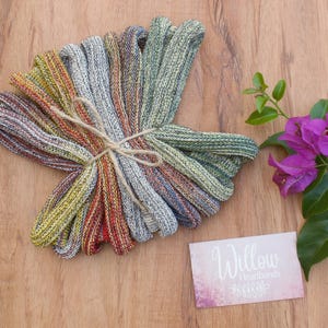 Puede incluir: Un conjunto de diademas multicolores atadas con hilo, en tonos rojos, amarillos, verdes y grises. Las diademas se muestran sobre una superficie de madera, con una tarjeta de visita "Willow Headbands" y una rama de flores moradas.