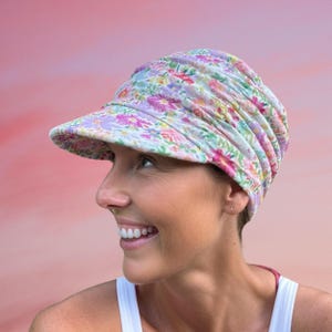以下が含まれることがあります： 柔らかくギャザーの入ったクラウンが特徴的な、花柄のバイザーハット。つばがあり、ピンク、紫、黄色の花が明るい背景に飾られています。帽子は人がかぶっています。