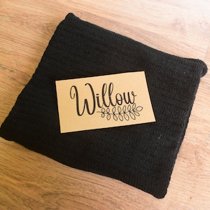 Könnte beinhalten: Ein gefalteter schwarzer Strickartikel mit einer rechteckigen Karte obenauf. Die Karte ist beige mit dem Namen "Willow" in Schreibschrift, mit einem dekorativen Blattdesign darunter. Der Artikel liegt auf einer hellbraunen Holzoberfläche.