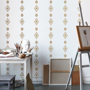 May include: A white room with a wooden table and two stools. The walls are decorated with a gold geometric pattern wallpaper. There is an easel with a blank canvas and a painting on it.