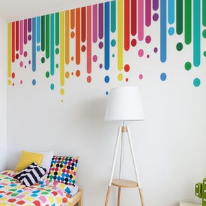 May include: A colorful bedroom with a bed, pillows, and a floor lamp. The bed has a multi-colored geometric patterned comforter. The wall is decorated with a rainbow-colored dripping paint design. A white lamp with wooden legs stands next to the bed.