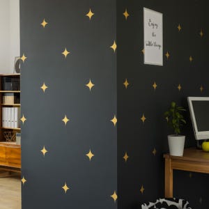 May include: Dark gray walls adorned with gold star decals. A framed print with the text "Enjoying the little things" hangs on the wall. A wooden desk with a computer and a potted plant is visible.