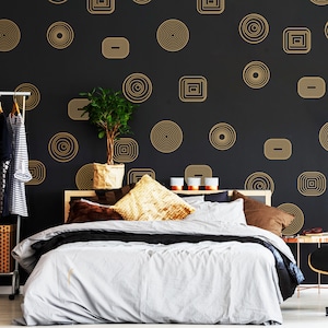 May include: A bedroom with a black wall featuring a gold geometric pattern of circles and squares. A bed with a gray duvet and brown pillows is in the foreground. A potted plant is on a shelf in the background.