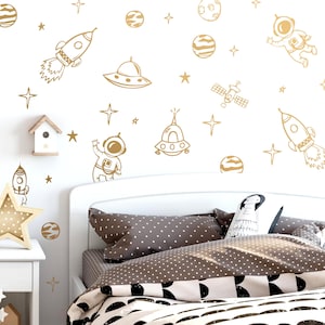May include: A bedroom scene with space-themed wall decals. The decals include rockets, planets, stars, astronauts, and UFOs in a gold color. The bed has a white frame, gray polka dot pillows, and a patterned comforter.