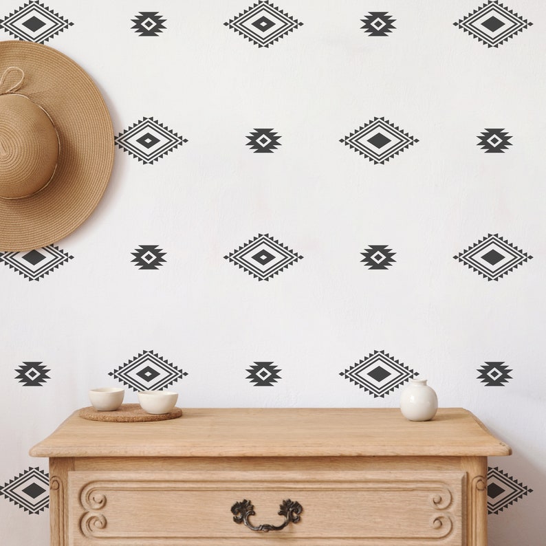 May include: A white wall with a repeating pattern of black diamond-shaped geometric designs. A wooden dresser with a drawer is in the foreground. A straw hat is on the wall to the left of the dresser.