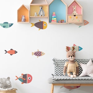 Op de afbeelding: Een witte muur met kleurrijke vis stickers en houten planken in de vorm van huizen. De planken zijn versierd met knuffels en speelgoed. Een grijs en wit chevron gepatroneerde bank met een knuffelhert staat voor de muur.