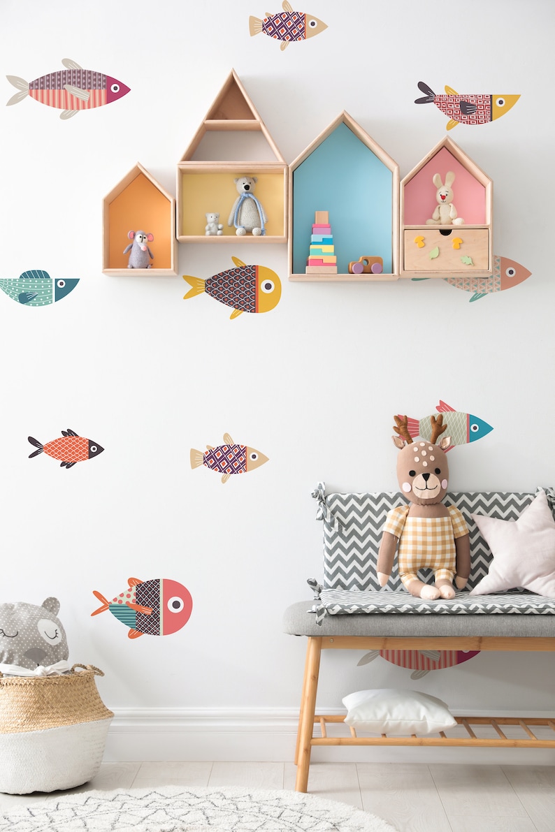 Op de afbeelding: Een witte muur met kleurrijke vis stickers en houten planken in de vorm van huizen. De planken zijn versierd met knuffels en speelgoed. Een grijs en wit chevron gepatroneerde bank met een knuffelhert staat voor de muur.