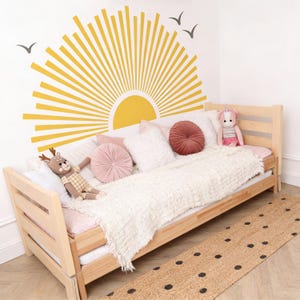May include: A light wood daybed is adorned with a white textured blanket and several pillows in shades of pink and white. A stuffed deer and unicorn are placed on the bed. A yellow sunburst design decorates the wall above the bed, with three black bird silhouettes.
