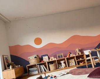 Może przedstawiać: Mural ścienny przedstawiający stylizowany krajobraz z warstwowymi wzgórzami w odcieniach ciemnego błękitu, fioletu i koralu, zwieńczony pełnym pomarańczowym słońcem. Scena jest namalowana na białej ścianie nad regałami z jasnego drewna z zabawkami.