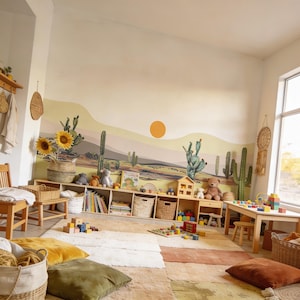 K&ouml;nnte beinhalten: Ein Wandbild mit einer W&uuml;stenlandschaft, gr&uuml;nen Kakteen, sanften H&uuml;geln in Gelb- und Beiget&ouml;nen sowie einer soliden orangefarbenen Sonne. Die Szene ist auf eine gro&szlig;e Wand in einem Raum mit h&ouml;lzernen Aufbewahrungsf&auml;chern, Spielzeug und Kissen gemalt.