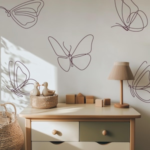May include: A light-colored dresser with two drawers, one white and one olive green, topped with a lamp and wooden blocks. The wall features line art butterfly designs. A wicker basket and two duck figurines are also present.
