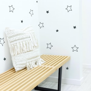 May include: A cream-colored macrame pillow with fringe rests on a light wood bench with black legs. The wall behind the bench is white with black star decals. The room has a modern, minimalist aesthetic.