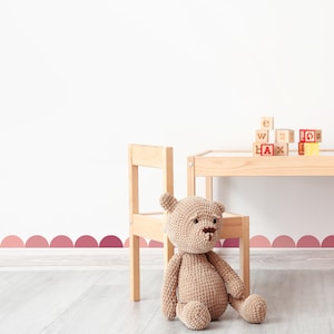 May include: A wooden table and chair set with a teddy bear sitting on the chair. The table has a stack of wooden blocks with letters on them. The wall has a pink scalloped border.