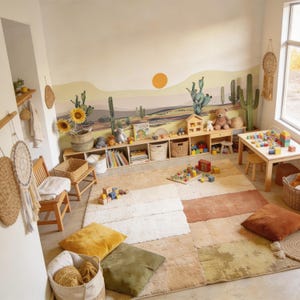 K&ouml;nnte beinhalten: Ein Kinderspielzimmer mit einem Wandbild im W&uuml;stenstil, das eine Sonne und Kakteen zeigt, einem langen Holzregal mit K&ouml;rben und Spielzeug, einem kleinen Holztisch mit Bausteinen, einem strukturierten Teppich sowie verschiedenen Bodenkissen und K&ouml;rben.