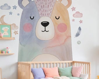 May include: A large, watercolor-style bear wall decal in pastel colors, with a matching crib and pillows. The bear is divided into sections of blue, green, and orange. Additional decals include stars, clouds, and flowers. A wooden crib is in the foreground.